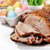 Half sliced Roasted Lamb Leg with carrot cake in the back (right corner). Left corner has Colorful easter themed eggs and barely visible bread.