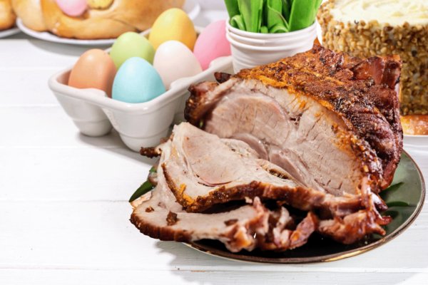 Half sliced Roasted Lamb Leg with carrot cake in the back (right corner). Left corner has Colorful easter themed eggs and barely visible bread.