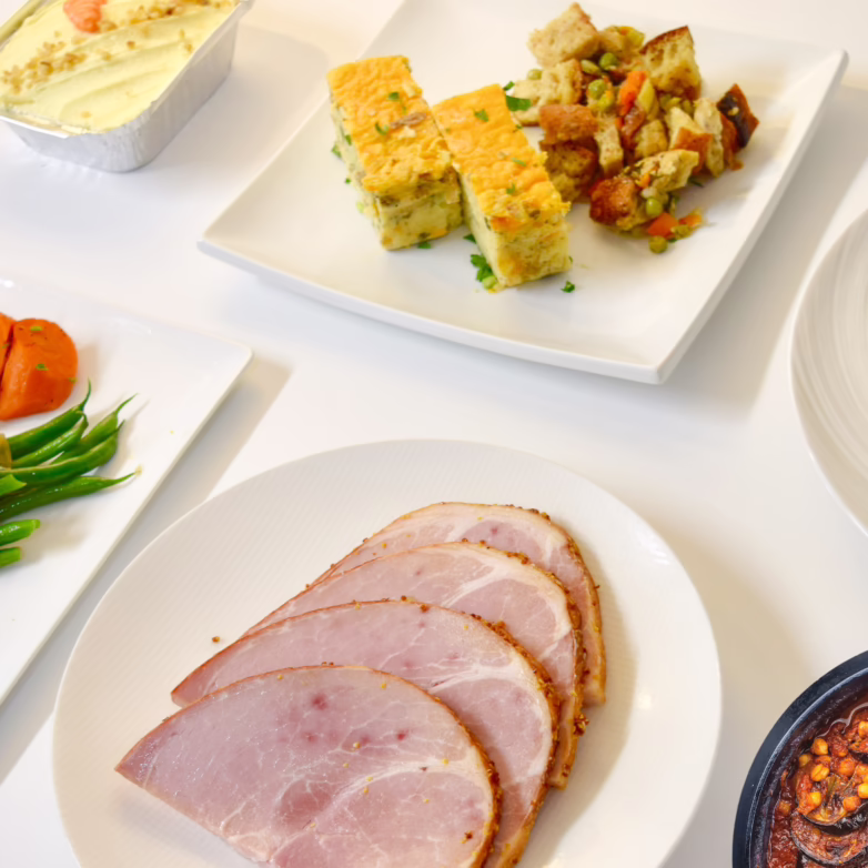 Plated on the table Easter dinner components including 4 slices of Ham, stuffing and potato casserole in the center. To the left beans with sauteed onions, carrots, and carrot cake. To the right carrot soup and vegetarian add on - Vegetable Moussaka.