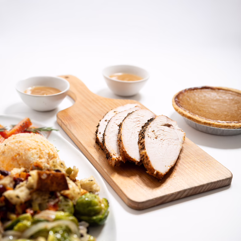 Sliced roast turkey breast on a wooden serving board, with bowls of gravy, a pumpkin pie, plate of yam and potato casserole with Brussels sprouts